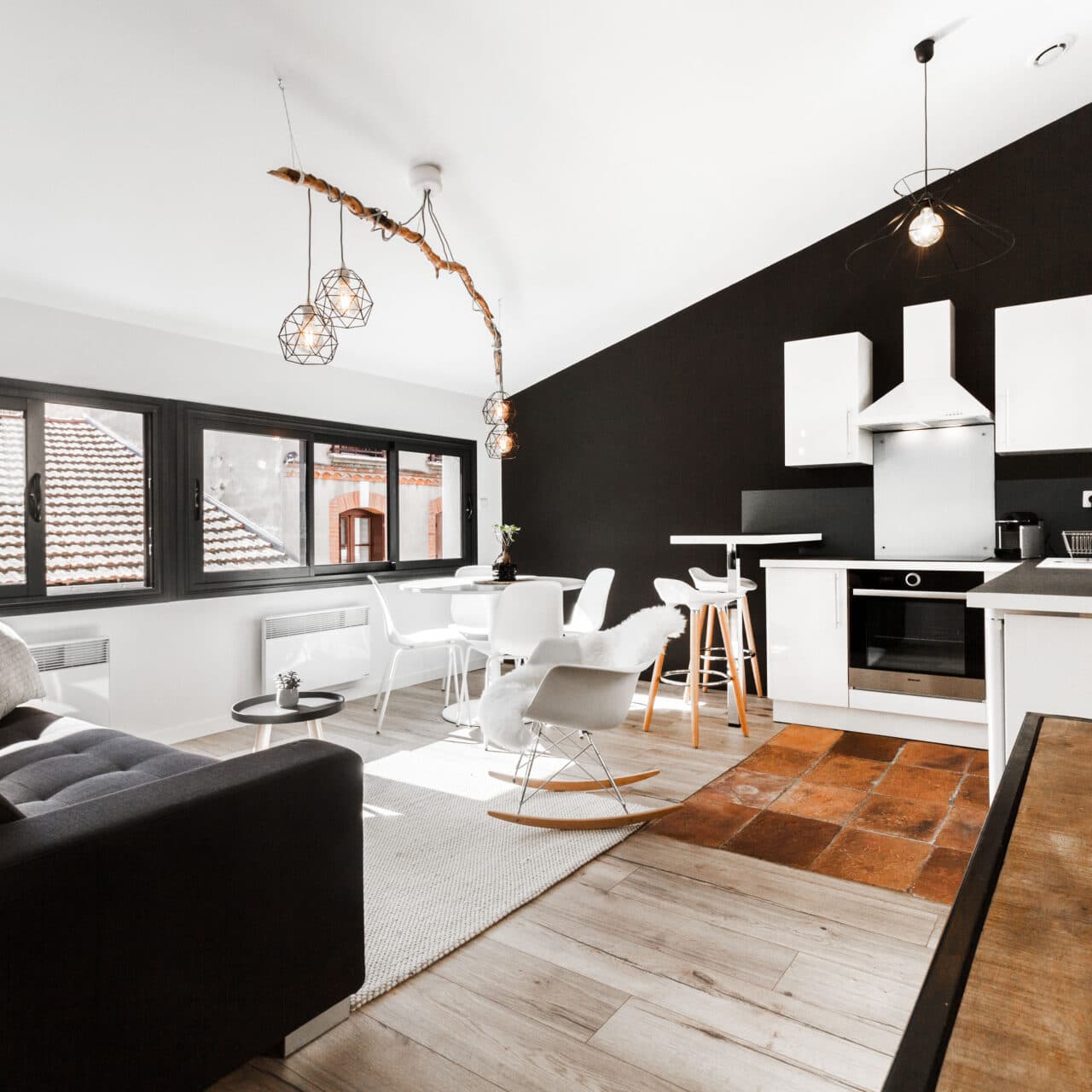 Séjour rénové ouvert sur la cuisine, mur noir mat et tomettes conservées au sol